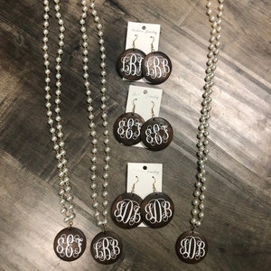 May include: A collection of fashion jewelry featuring pearl necklaces and matching earrings. The earrings and pendant are dark brown with white monogram letters. The necklaces are long with small, round, white pearls.