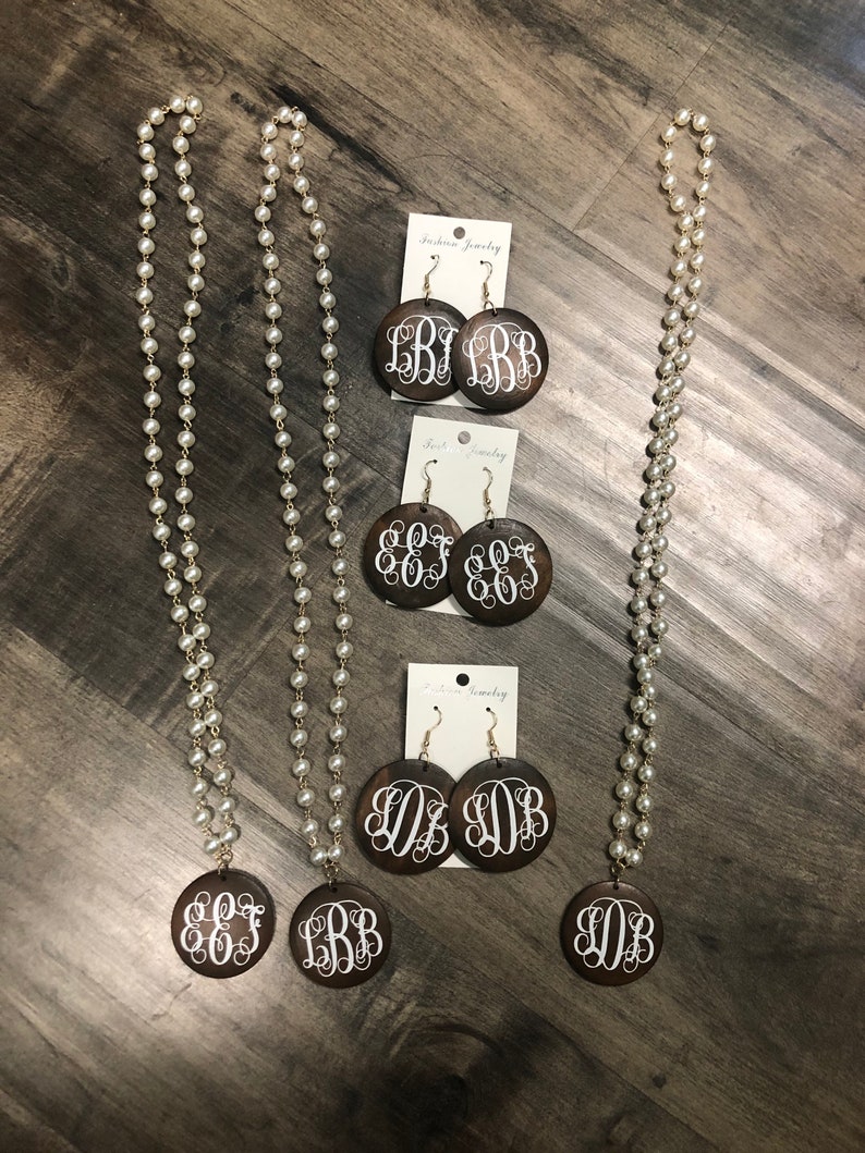 May include: A collection of fashion jewelry featuring pearl necklaces and matching earrings. The earrings and pendant are dark brown with white monogram letters. The necklaces are long with small, round, white pearls.