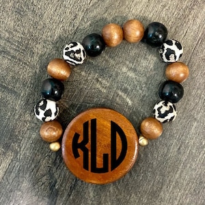 Può includere: Braccialetto di perline di legno con un grande ciondolo rotondo in legno con le iniziali "KLD" in nero. Il braccialetto include perline nere, marroni e con stampa leopardata. Un accessorio elegante per l'uso quotidiano.