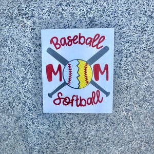 May include: A white square sign with the words "Baseball Mom Softball" in red script. A baseball and two crossed bats are in the center. The baseball is white, yellow, and red. The background is a textured silver.
