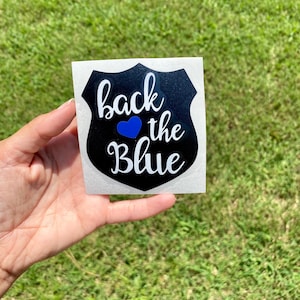 Può includere: Un adesivo bianco con un disegno di distintivo della polizia nero. Il distintivo riporta le parole "back the Blue" in corsivo bianco, con un cuore blu che sostituisce la parola "of". L'adesivo è tenuto su uno sfondo erboso verde.