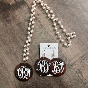 May include: A pearl necklace and matching earrings set. The necklace features faux pearls strung on a gold-tone chain. The earrings and pendant are dark brown wood with a white monogram design. The earrings hang from gold-tone hooks.