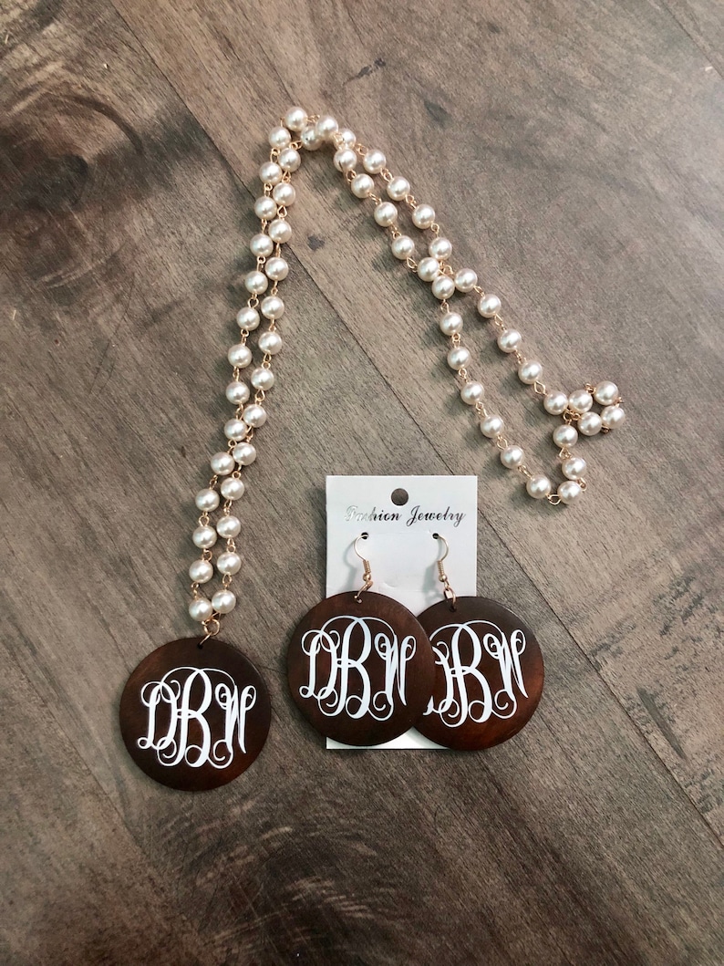 May include: A pearl necklace and matching earrings set. The necklace features faux pearls strung on a gold-tone chain. The earrings and pendant are dark brown wood with a white monogram design. The earrings hang from gold-tone hooks.