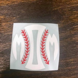 Pu&ograve; includere: Adesivo bianco a tema baseball con un design a cucitura rossa. L'adesivo presenta una forma di baseball con la lettera "M" su ogni lato. L'adesivo &egrave; su una superficie di legno.