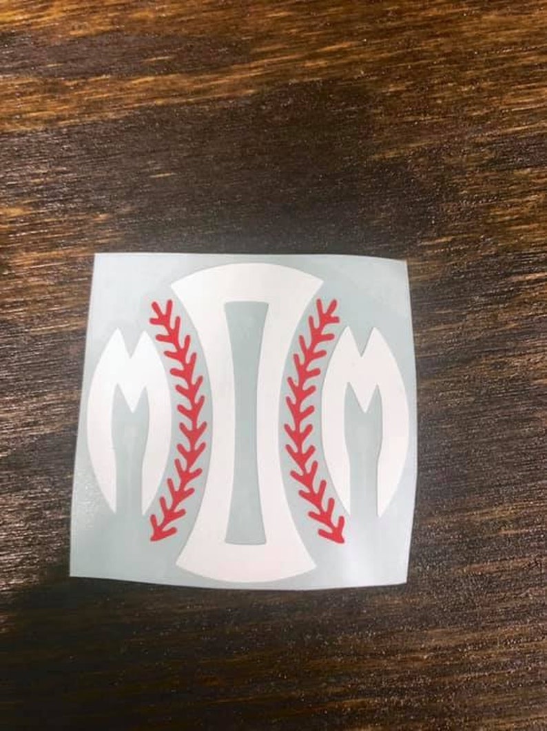 Pu&ograve; includere: Adesivo bianco a tema baseball con un design a cucitura rossa. L'adesivo presenta una forma di baseball con la lettera "M" su ogni lato. L'adesivo &egrave; su una superficie di legno.