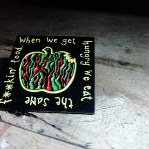 Könnte beinhalten: Schwarzer Emaille-Pin mit gelber Umrandung. Der Pin zeigt einen roten, grünen und schwarzen Apfel mit einem Bissen. Der Text "When we get hungry we eat the same f***kin food." ist um den Apfel herum geschrieben.