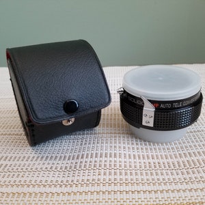 May include: A black leather case with a silver button and a black and silver teleconverter lens with the text "Soligor MP Auto Tele Converter" on the side.