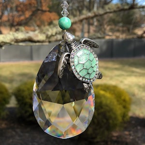 May include: A decorative suncatcher featuring a faceted crystal teardrop pendant. A silver-toned turtle charm with a mint green shell and crystal accents is attached. The suncatcher is suspended by a strand of clear crystals and green beads.