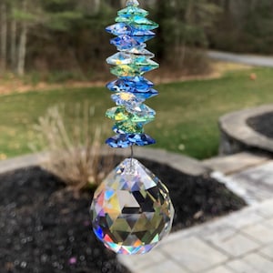 May include: A hanging crystal suncatcher with a large faceted crystal ball at the bottom and a series of blue, green, and clear crystal accents above. The suncatcher is suspended by a thin wire, reflecting light.