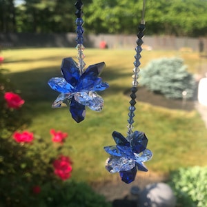 May include: Two hanging crystal flower ornaments. Each ornament features a faceted blue and clear crystal flower suspended from a string of beads. The background is a blurred outdoor scene with greenery and sunlight.