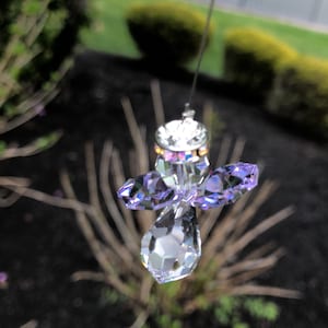 May include: A crystal angel suncatcher with iridescent purple wings and a clear crystal body. The suncatcher is hanging from a silver string.
