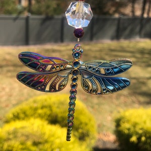 May include: A decorative dragonfly suncatcher with iridescent wings in shades of blue, purple, and gold. The dragonfly is suspended from a clear faceted crystal and purple beads, designed for hanging. This garden decor item adds a touch of whimsy.