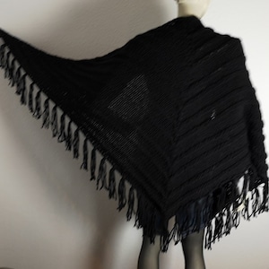 Könnte beinhalten: Ein schwarzer Strickschal mit Fransen. Der Schal ist über einer Schaufensterpuppe drapiert und zeigt seine dreieckige Form und das strukturierte Strickdesign. Die Fransen hängen an den Kanten.