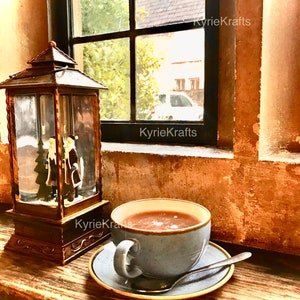 May include: A blue ceramic mug filled with a warm drink sits on a wooden surface next to a decorative lantern with a snow globe inside. The mug is on a blue and white saucer with a silver spoon. The window behind the mug is open and shows a view of a building and trees.