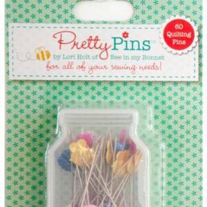 Op de afbeelding: Een doorzichtige plastic pot gevuld met 60 kleurrijke naalden voor quilten. De pot heeft een wit deksel en is gelabeld "Pretty Pins" door Lori Holt of Bee in my Bonnet. Het label vermeldt ook "voor al uw naaibehoeften!"
