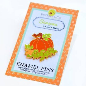 May include: Gold enamel pin featuring a pumpkin with green leaves. The pin is on a card that says "Fig Tree & Co. Seasons Collection" and "Enamel Pins" with the company website and Instagram handle.