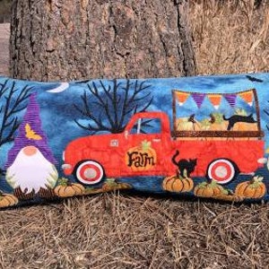 Könnte beinhalten: Ein langes, rechteckiges Kissen mit Halloween-Motiv. Das Kissen zeigt einen roten LKW mit dem Wort "Farm" an der Seite, der Kürbisse und eine schwarze Katze transportiert. Zwei Gnome mit spitzen Hüten und weißen Bärten befinden sich auf beiden Seiten des LKWs. Der Hintergrund ist dunkelblau mit weißen Wolken und Bäumen.