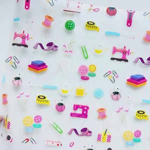 May include: A translucent sheet featuring a repeating pattern of colorful sewing supplies, including pink sewing machines, yellow measuring tapes, green and yellow buttons, spools of thread, fabric stacks, safety pins, and pincushions.