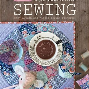 Puede incluir: Un libro titulado "Hot Chocolate Sewing: Cozy Autumn and Winter Sewing Projects" de Tilda. La portada presenta una colcha de retazos con una taza de chocolate caliente, un pequeño animal de peluche y carretes de hilo.