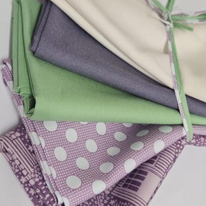 Puede incluir: Una pila de piezas de tela dobladas en varios colores y patrones, incluyendo crema sólido, gris sólido, verde claro sólido y tela morada con lunares blancos y diseños florales geométricos, todo atado con una tira de tela verde delgada.
