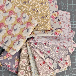 Könnte beinhalten: Eine Sammlung gefalteter Stoffquadrate mit verschiedenen floralen Mustern. Die Stoffe weisen eine Reihe von Farben auf, darunter Rosa, Gelb, Blau und Grau. Die Designs umfassen Blumen, Blätter und Beeren. Die Stoffe sind auf einer grauen Schneidematte angeordnet.
