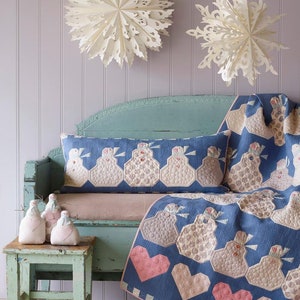 May include: A blue quilt and cushion set featuring a snowman and heart pattern. The quilt is draped over a distressed blue bench. Paper snowflakes are mounted on the wall. Three snowman figurines sit on a small, matching stool, all in a pastel colour scheme.
