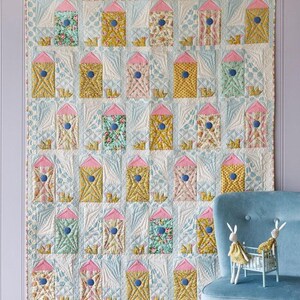 May include: A patchwork quilt featuring a repeating design of houses in various colours and patterns. Each house has a pink roof and blue detailing. The quilt is displayed against a pale lilac wall.