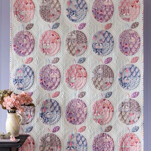 May include: A large, quilted wall hanging with a repeating apple design. Each apple is a different colour and pattern, including pink, blue, purple, and brown. The quilt is hung against a light blue background.