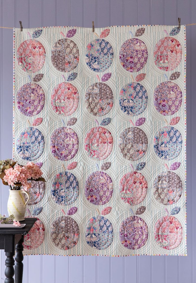 May include: A large, quilted wall hanging with a repeating apple design. Each apple is a different colour and pattern, including pink, blue, purple, and brown. The quilt is hung against a light blue background.