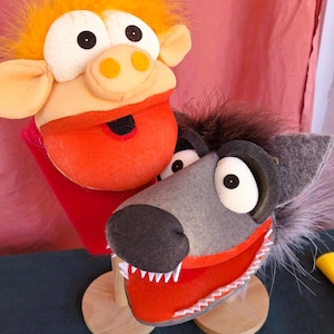 May include: Two handmade puppet heads. One is a pig with orange lips and a yellow nose, and the other is a gray wolf with white teeth and an orange mouth. Both puppets have large, expressive eyes and are mounted on wooden bases.