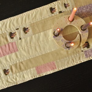 May include: A cream, tan, and pink patchwork table runner with a decorative wooden candle holder. The candle holder has lit candles. The runner features repeating patterns of a small house and floral motifs.