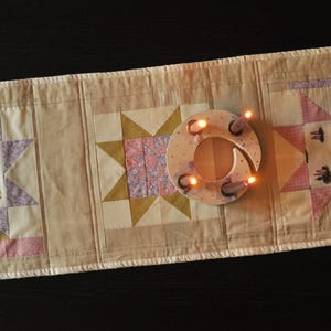 May include: A cream-coloured quilted table runner with star-shaped patchwork designs in pink, yellow, and lavender. A circular candle holder with lit candles sits on the runner, creating a warm ambiance. The runner is approximately 152 cm long.