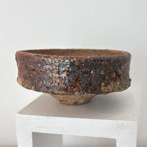 May include: A handcrafted, rustic ceramic bowl with a wide, textured rim. The bowl features a dark brown exterior with lighter brown and beige tones, and a slightly flared base. The bowl is displayed on a white, square pedestal.