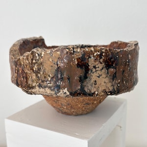 May include: A handcrafted, textured ceramic bowl with a wide, irregular rim. The bowl features a blend of brown, tan, and black hues, creating a rustic aesthetic. The bowl is elevated on a white, square pedestal, showcasing its unique design.