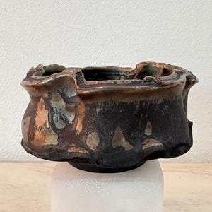 May include: A ceramic bowl with an irregular, wavy rim and a dark, mottled glaze. The surface features a blend of deep black, metallic copper, and muted blue tones, creating a textured, uneven appearance with a wide, open top and a narrow base.