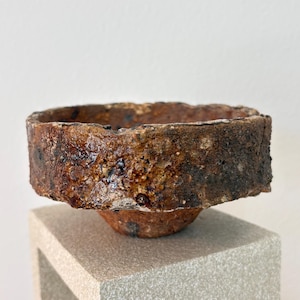May include: A handcrafted, rustic ceramic bowl with a rough, textured rim. The bowl is a warm brown color with darker, mottled areas, and sits atop a square, light gray pedestal. The bowl's design suggests a focus on natural aesthetics.