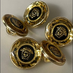 May include: Five gold-toned buttons with a black center featuring a gold-colored design. The buttons have a raised, textured rim.
