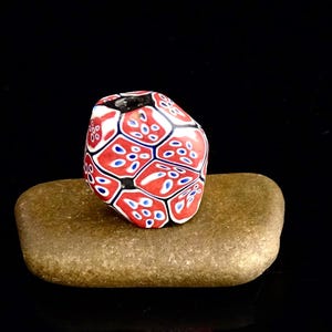 Può includere: Un perno di vetro rosso, bianco e blu con un motivo geometrico. Il perno è seduto su una roccia marrone.