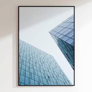 May include: Framed architectural print featuring two modern glass skyscrapers against a pale sky. The buildings have many windows, reflecting the sky and creating a blue and gray color palette. The black frame adds a clean, contemporary look.