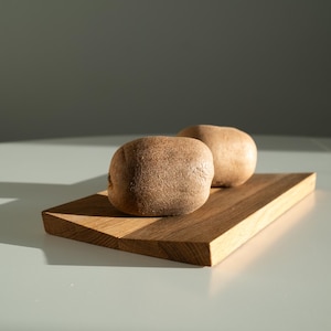 May include: Two whole kiwi fruits with brown, fuzzy skin resting on a rectangular wooden cutting board. The board features a visible wood grain pattern and light brown tones, positioned under direct light creating a sharp shadow.