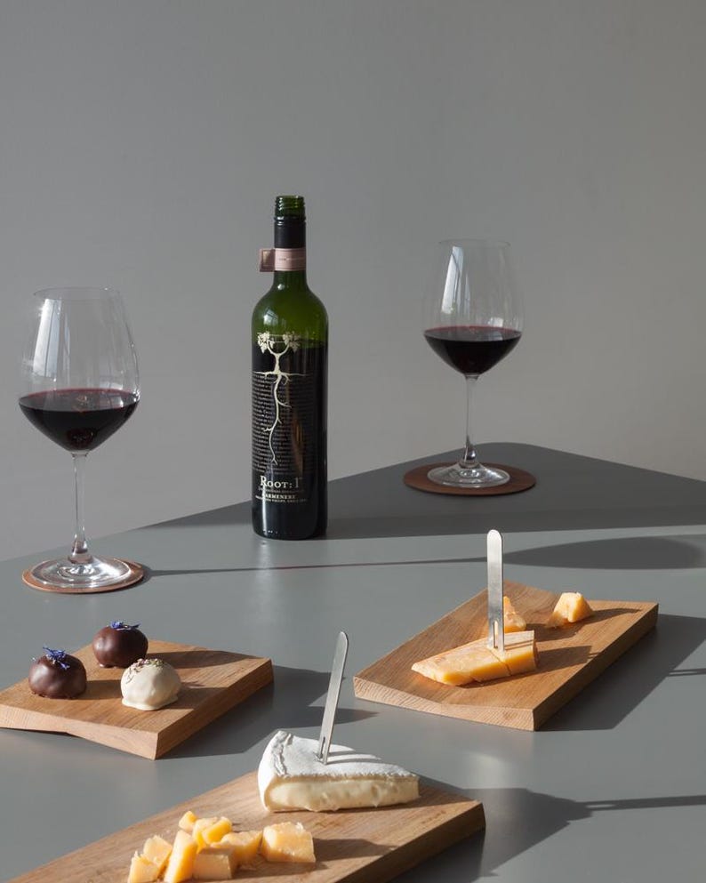 May include: A dark green wine bottle labeled Root: 1, two stemmed glasses filled with red wine, three rectangular wooden serving boards, two silver metal cheese forks, assorted cheese wedges, and three chocolate truffles with floral garnishes.