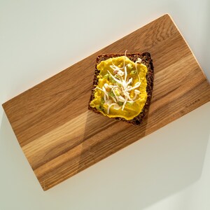 May include: A rectangular wooden serving board with a natural grain pattern and the word minumo engraved on the right side. A slice of dark brown bread topped with yellow hummus and white bean sprouts is placed on the board's surface.