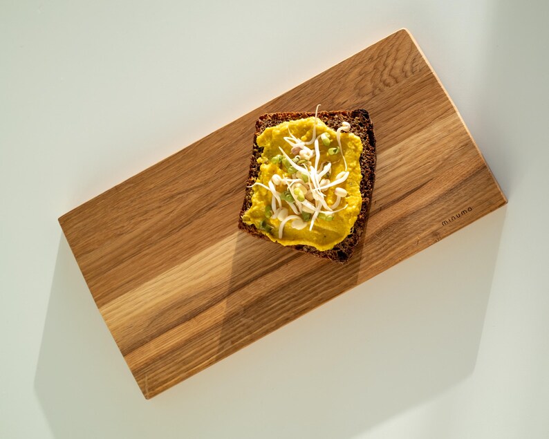 May include: A rectangular wooden serving board with a natural grain pattern and the word minumo engraved on the right side. A slice of dark brown bread topped with yellow hummus and white bean sprouts is placed on the board's surface.