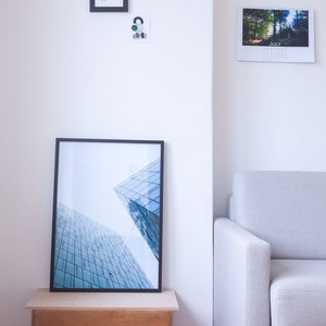 A black framed photograph of a modern building with a blue glass facade. The photograph is displayed on a wooden shelf with a white wall in the background. A white sofa is in the corner of the room.