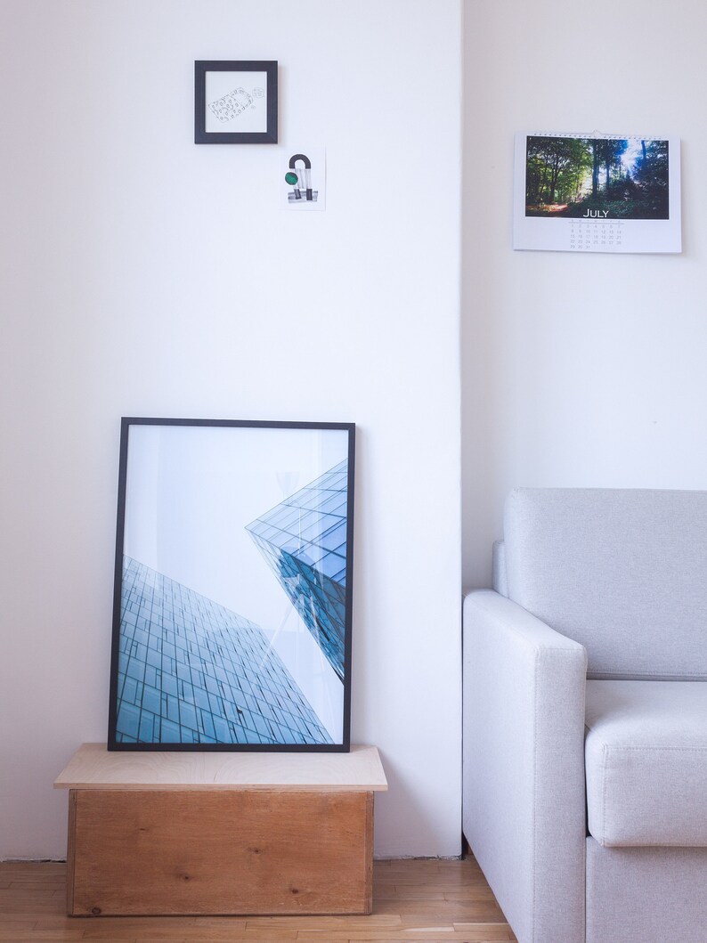 A black framed photograph of a modern building with a blue glass facade. The photograph is displayed on a wooden shelf with a white wall in the background. A white sofa is in the corner of the room.