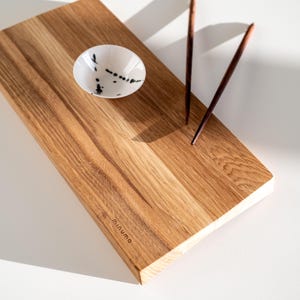 May include: A rectangular wooden serving board with a natural grain finish and the word minumo embossed on the corner. It features a small white ceramic bowl with black speckles and a pair of dark brown wooden chopsticks resting on the surface.