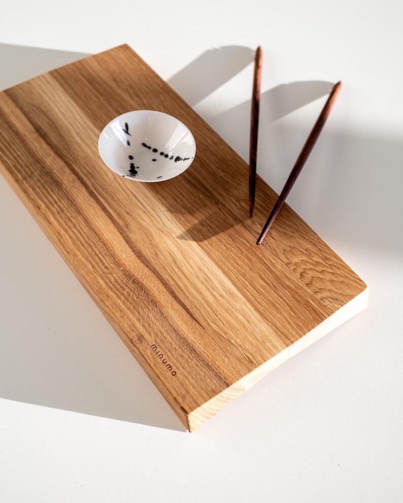 May include: A rectangular wooden serving board with a natural grain finish and the word minumo embossed on the corner. It features a small white ceramic bowl with black speckles and a pair of dark brown wooden chopsticks resting on the surface.