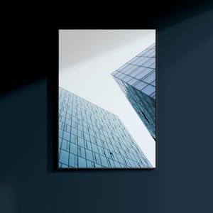 May include: Framed architectural photograph featuring two modern glass skyscrapers against a pale sky. The buildings have many windows, reflecting the sky and creating a geometric pattern. The frame is black, and the wall is a dark blue.