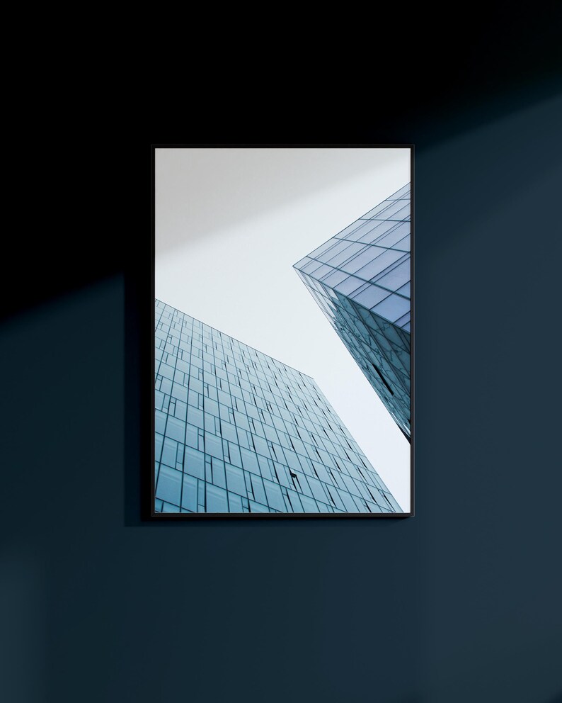 May include: Framed architectural photograph featuring two modern glass skyscrapers against a pale sky. The buildings have many windows, reflecting the sky and creating a geometric pattern. The frame is black, and the wall is a dark blue.