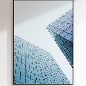 May include: Framed architectural print featuring two modern buildings with blue-tinted glass facades against a white sky. The buildings have a geometric design with numerous windows, creating a contemporary aesthetic.